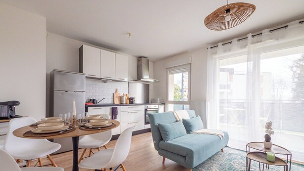 Eine moderne Küche mit weißen Schränken, ein Essbereich mit rundem Tisch und Stühlen, ein blaues Sofa und ein großes Fenster mit Blick auf Gebäude.