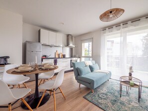 A modern kitchen with white cabinets, a dining area with a round table and chairs, a blue sofa, and a large window with a view of buildings. - Le Cocon de Saint Priest - T2 Balcony & Terrace (Saint-Priest)