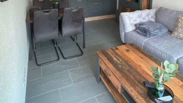 A modern kitchen with grey cabinets, a dining area with a table and chairs, a grey sofa with a blanket, and a wooden coffee table with a vase and plant. - Vacation apartment Pingo 1 (Ahrbrück)