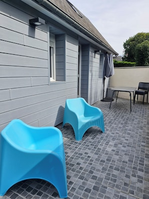 A patio area with a tiled floor, two blue plastic chairs, a small table, and a window. - 3★ House, Les 4 As, exceptional location 800 m from the sea, sleeps 6. (Veulettes-sur-Mer)