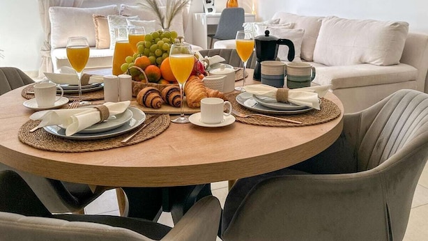 Un comedor con una mesa redonda dispuesta para el desayuno, que incluye frutas, croissants y bebidas.