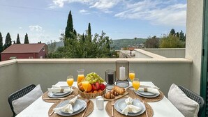 Un balcón con una mesa dispuesta para el desayuno, que incluye frutas, pan y bebidas, con vistas a un paisaje pintoresco con árboles y un edificio distante.