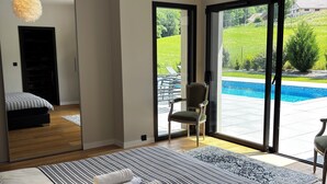 Une chambre moderne avec un grand lit, un luminaire suspendu et une vue sur une piscine, visible à travers une porte-fenêtre coulissante.
