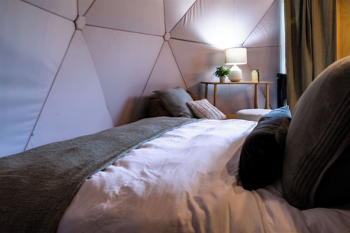 Ein modernes Hotelzimmer mit einem Kopfende im geometrischen Muster, einem ordentlich bezogenen Bett mit einer grauen Tagesdecke, einem kleinen Nachttisch mit Lampe und Pflanze sowie einem Blick ins Freie durch ein Fenster mit Vorhängen.