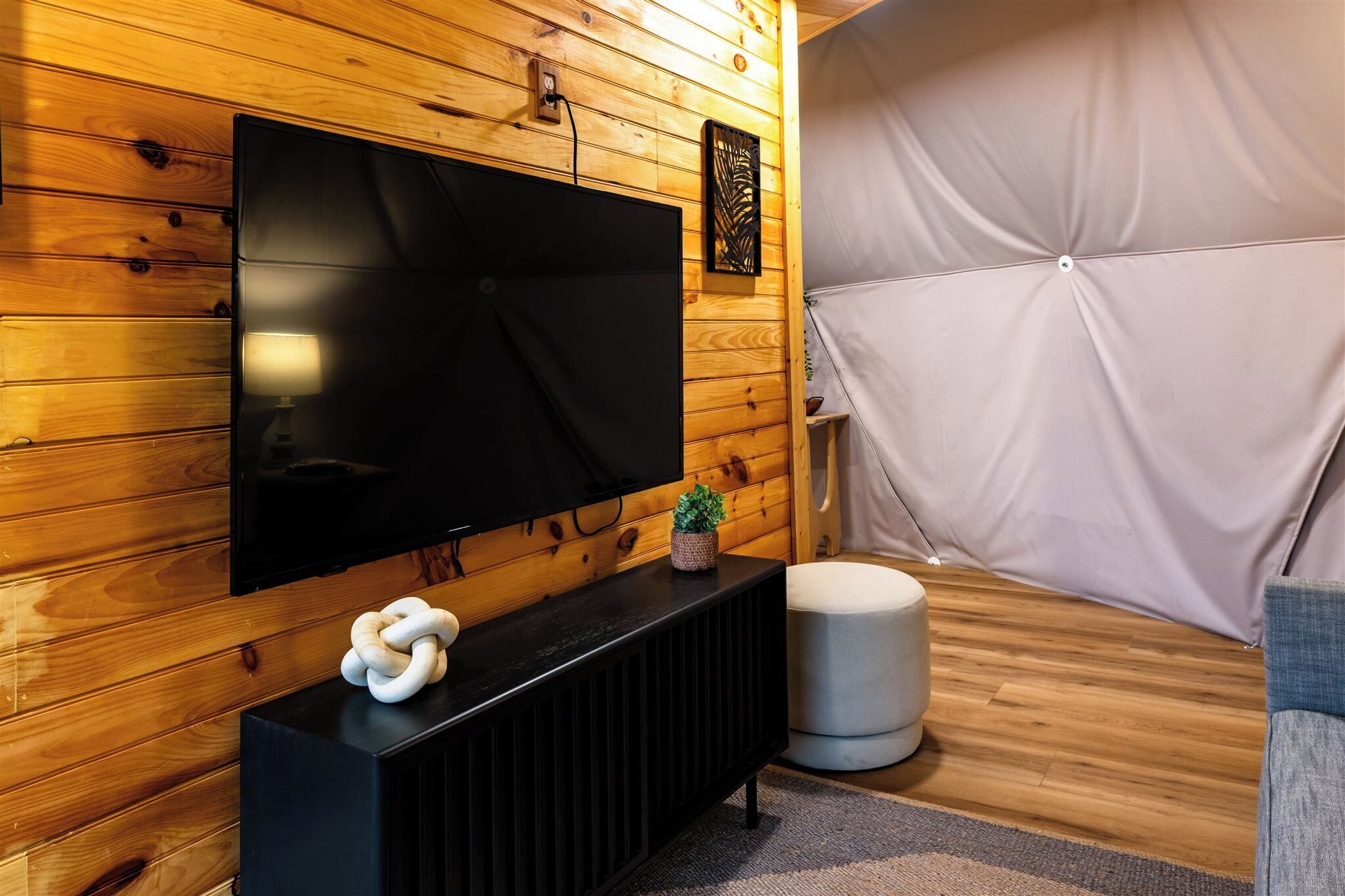 A modern room with a large flat-screen TV mounted on a wooden wall, a black console table with a decorative item, a round ottoman, and a cushioned seating area.