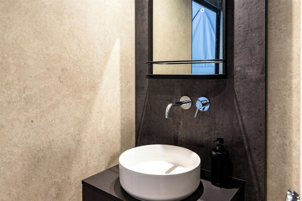 A modern bathroom with a round white sink, a black soap dispenser, and a mirror with a black frame.