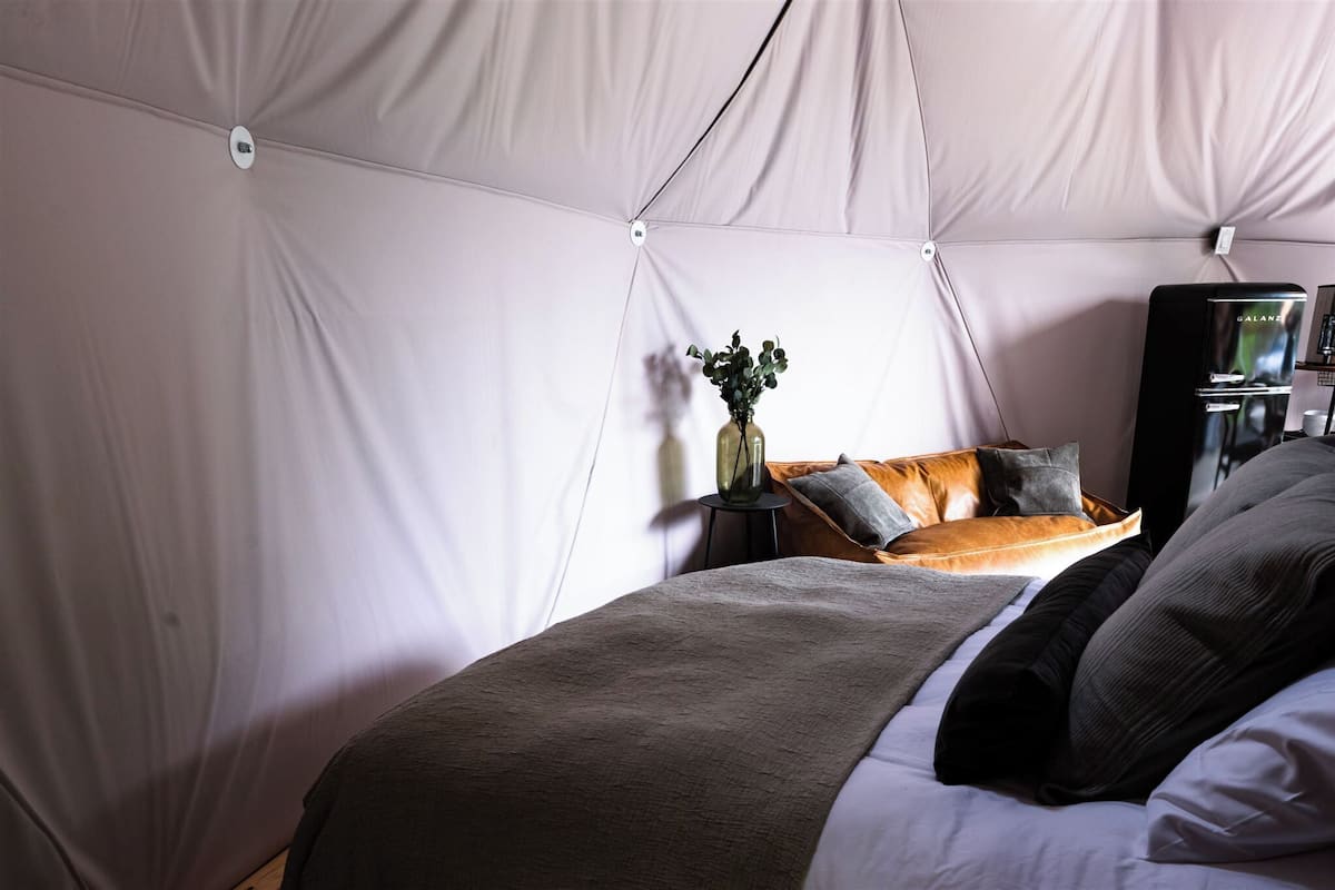 Ein Zelttraum mit Bett, Kissen, einem kleinen Tisch mit einer Vase und einem schwarzen Gerät.