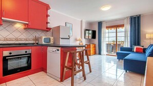 A compact kitchen with red cabinets, a built-in oven, and a small island with stools. A dining area with a blue sofa and a TV is visible. - Garden city with ocean view&heated pool (Costa Adeje)