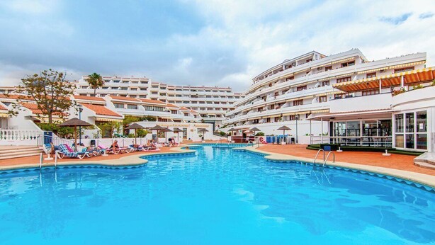 A swimming pool surrounded by multi-story hotel buildings with balconies, a covered seating area, and a clear sky. - Garden city with ocean view&heated pool (Costa Adeje)
