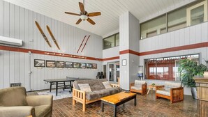 Ein großzügiger Loungebereich mit hohen Decken, Deckenventilator aus Holz und Holzboden. Es gibt verschiedene Sitzmöglichkeiten, darunter eine Couch, Stühle und ein kleiner Tisch. Die Wände sind mit gerahmten Bildern geschmückt, und ein großes Fenster sorgt für natürliches Licht.