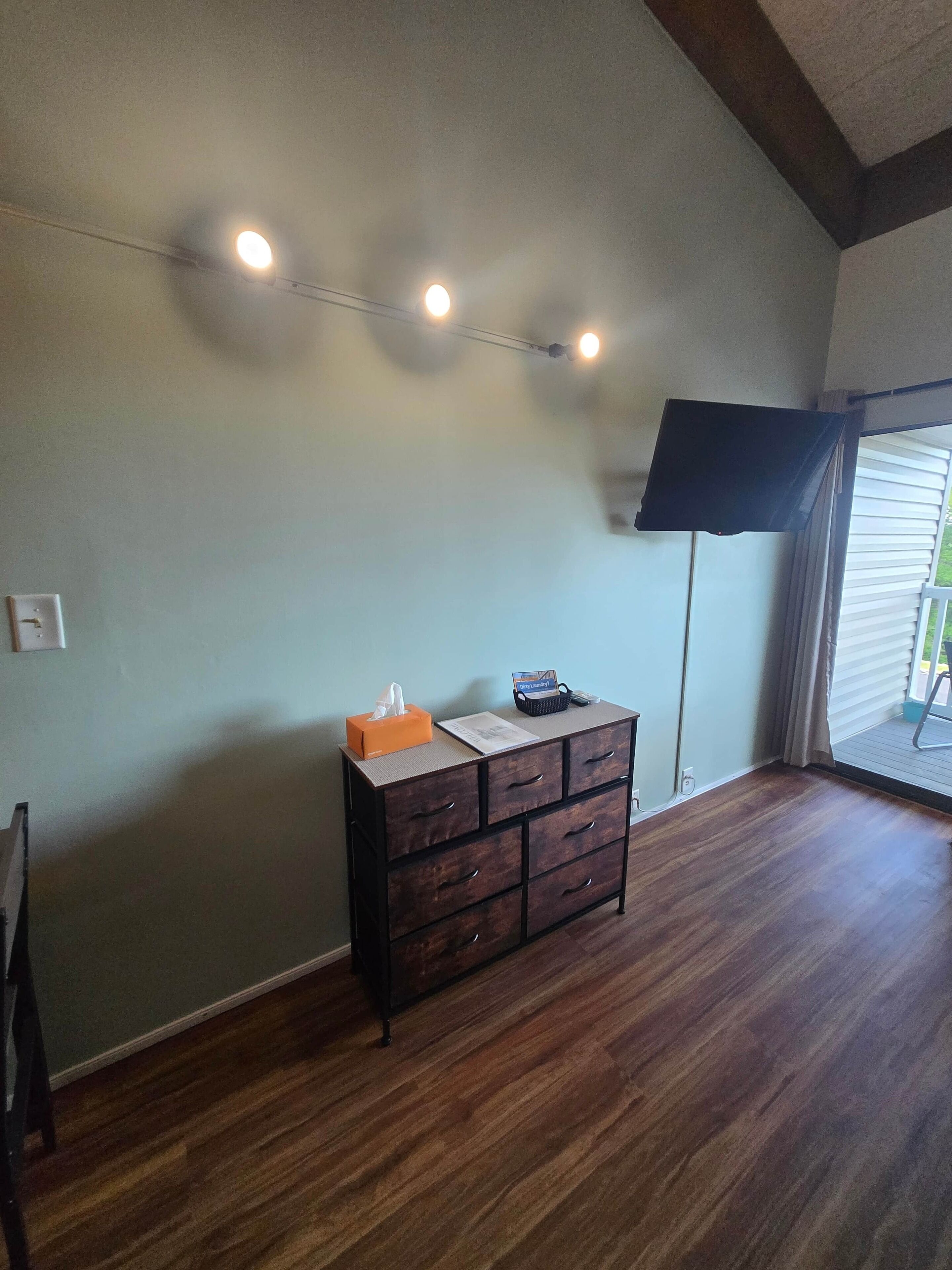 Quarto com cômoda de madeira, televisão de tela plana na parede e janela com cortinas.