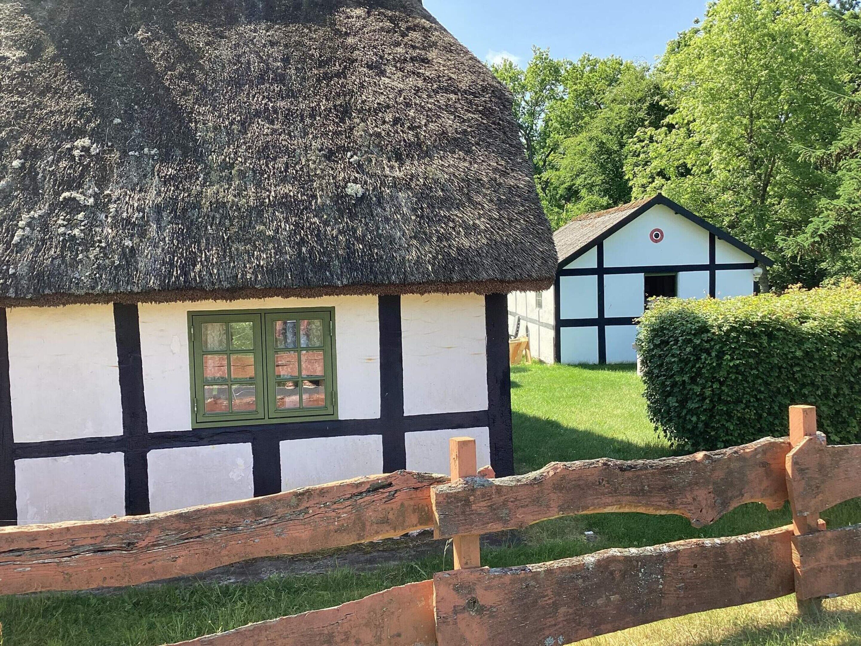 A traditional thatched-roof cottage with a white and black half-timbered facade, a green window, and a wooden fence in the foreground.