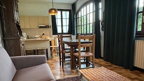 Una sala de estar con sofá, una mesa de comedor con sillas, un reposapiés de mimbre y una cocina al fondo.