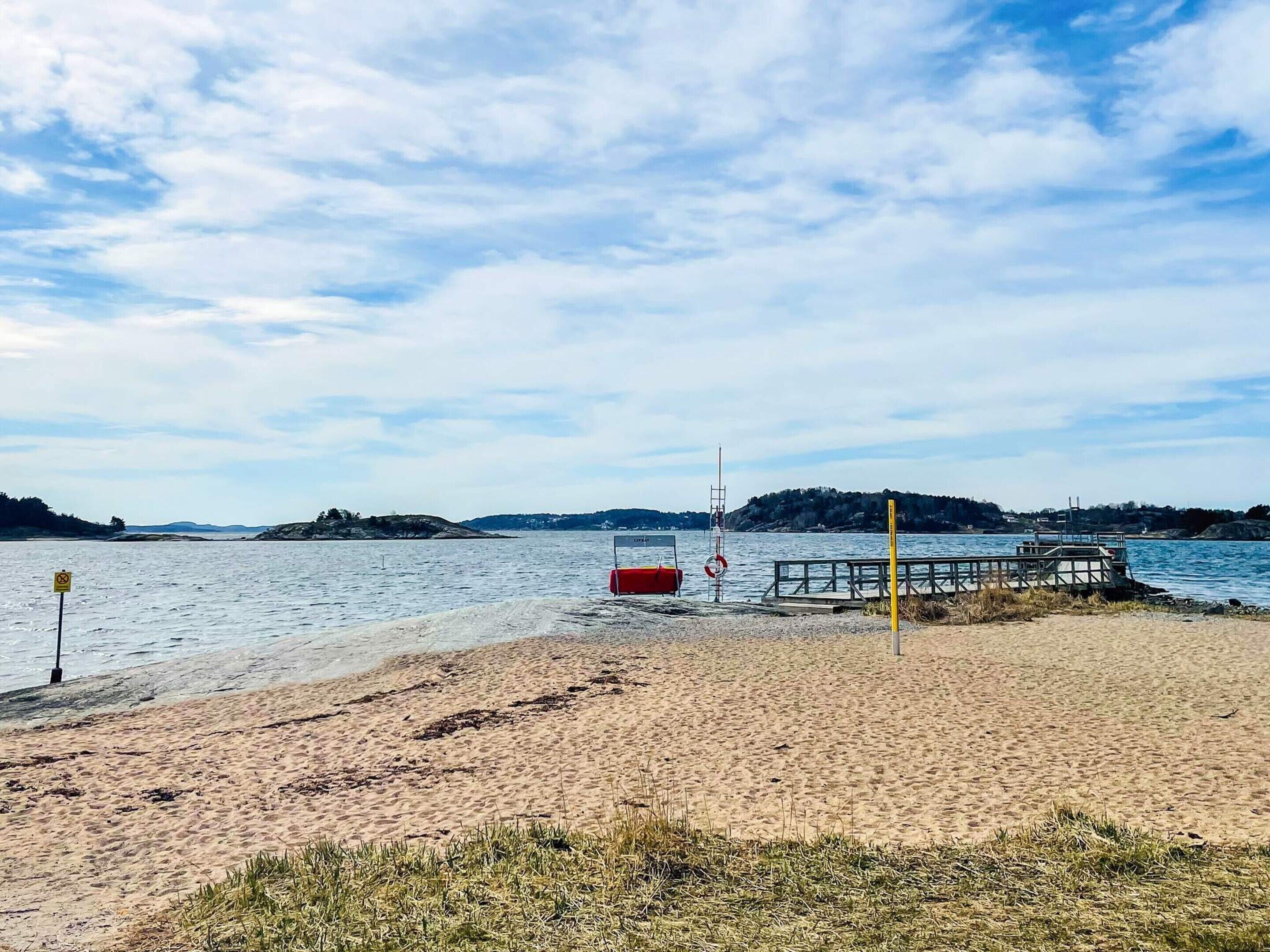 A beach with a wooden pier extending into calm water, a lifebuoy, and a no swimming sign.