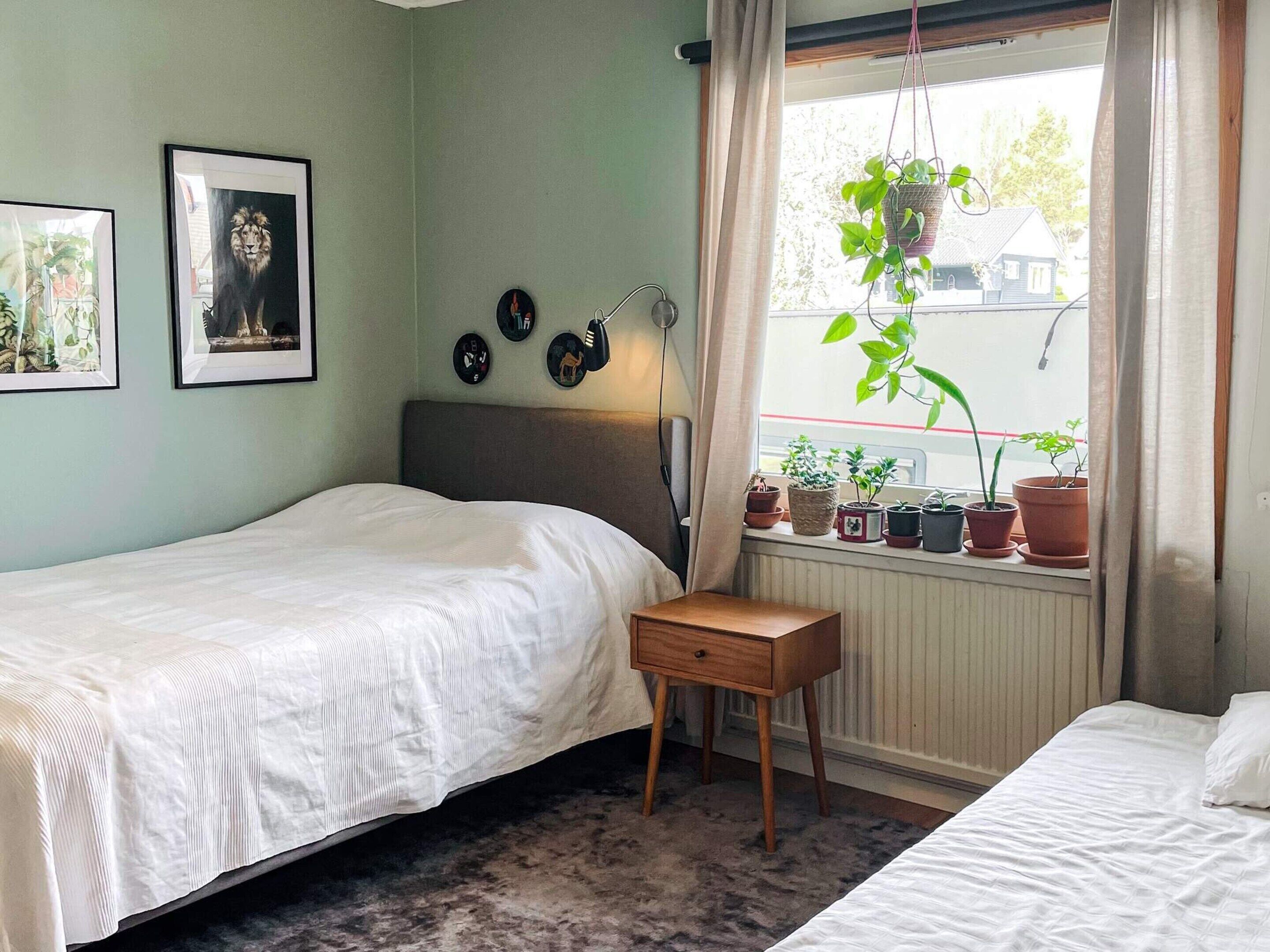 Een slaapkamer met een bed, een nachtkastje, een raam met gordijnen en diverse potplanten op de vensterbank.
