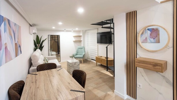 A modern living room with a wooden dining table, a sofa, and a light blue armchair. There is a TV mounted on the wall and a small side table with a vase.