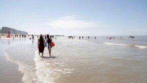Escena de playa con gente caminando por la orilla y un muelle al fondo.