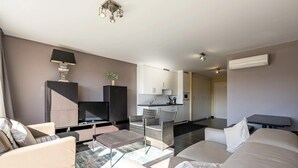 Una sala moderna con sofá, mesa de centro y televisor. Se observa una zona de cocina con armarios y una mesa de comedor.