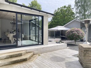 Uma casa moderna com porta de vidro que dá para uma sala de jantar, um pátio com móveis de exterior e um jardim com arbusto florido.