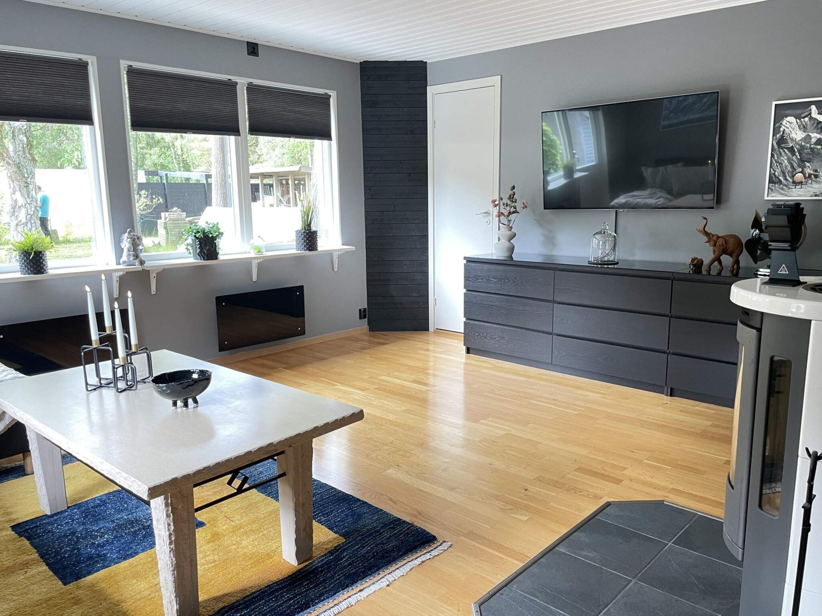 A modern living room with a wooden floor, a dining table with candles, a fireplace, a large TV, and a kitchen area with dark cabinetry.