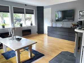Una sala moderna con suelo de madera, una mesa de comedor con velas, una chimenea, un televisor grande y una zona de cocina con armarios oscuros.