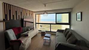A modern living room with a large window, a flat-screen TV, and a view of the beach.
