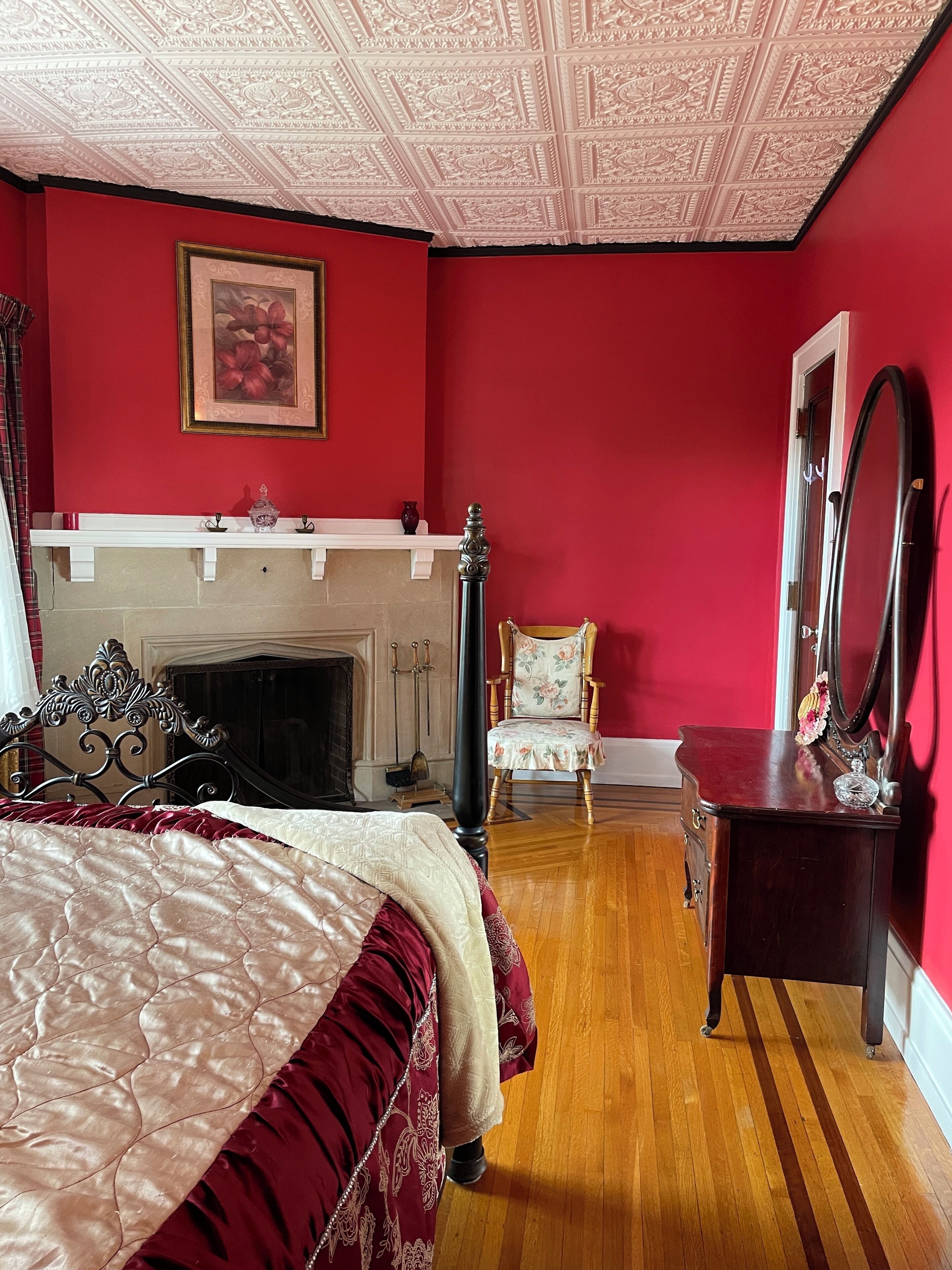 Une chambre avec un mur rouge, une cheminée, un lit recouvert d’une couverture rouge et blanche, une chaise et une commode avec un miroir.