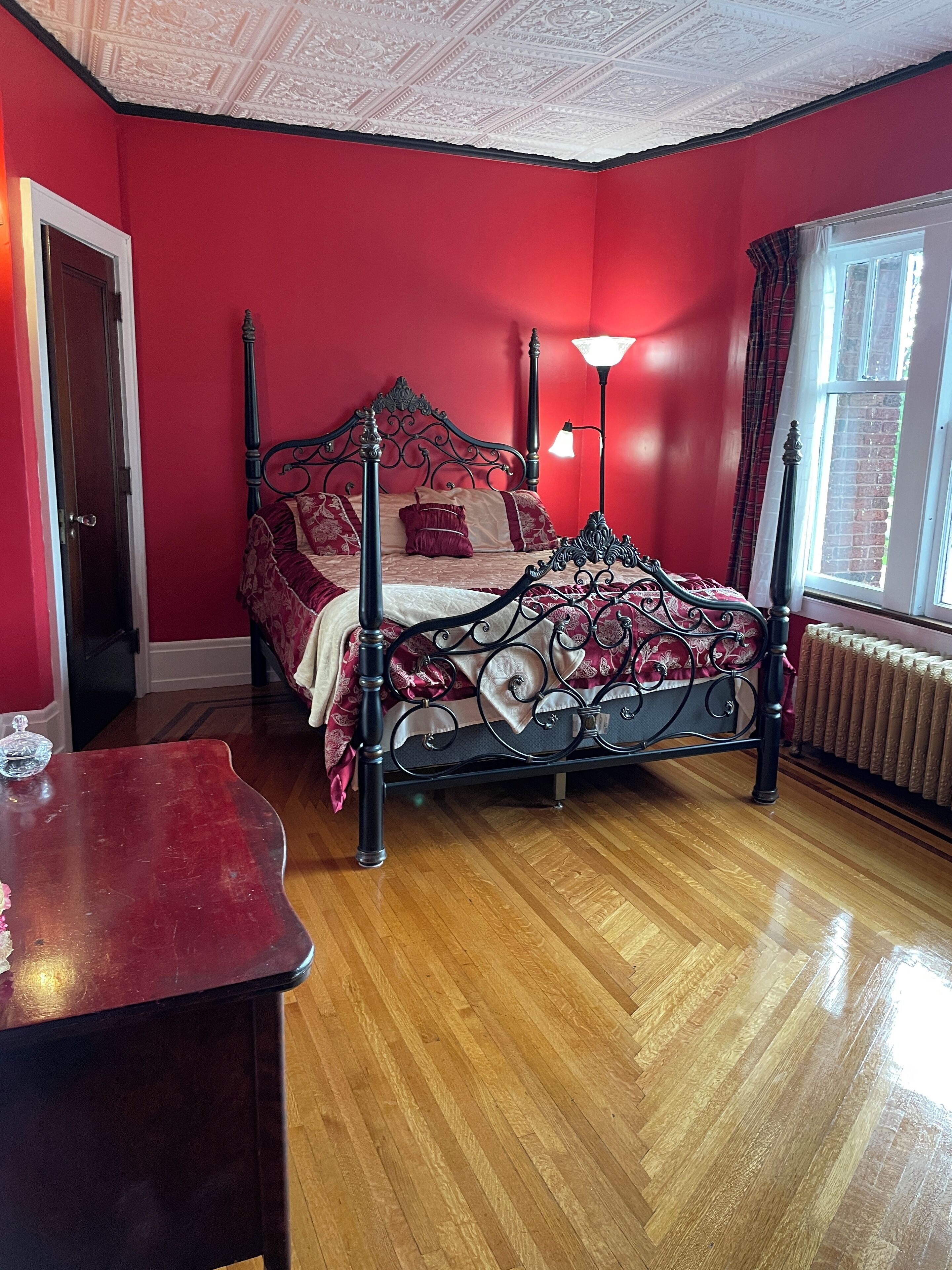 Une chambre à coucher avec un lit à baldaquin, un plancher en bois, un mur rouge, une fenêtre avec des rideaux et un radiateur.