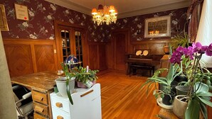 Une pièce avec un plancher en bois, une commode blanche, un bureau avec une chaise, un piano et diverses plantes en pot.