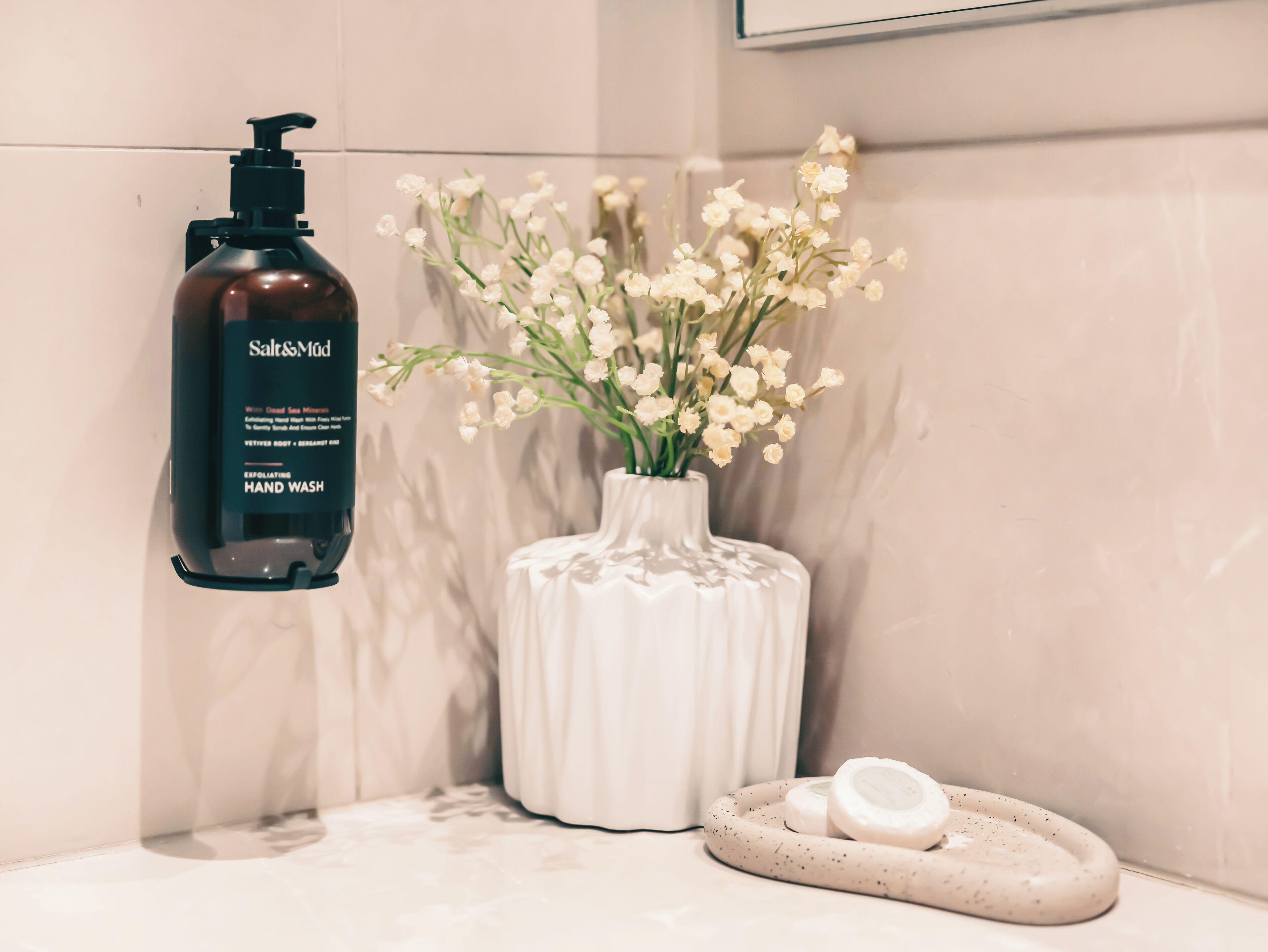 A bathroom with a hand soap dispenser, a vase with flowers, and a soap dish.