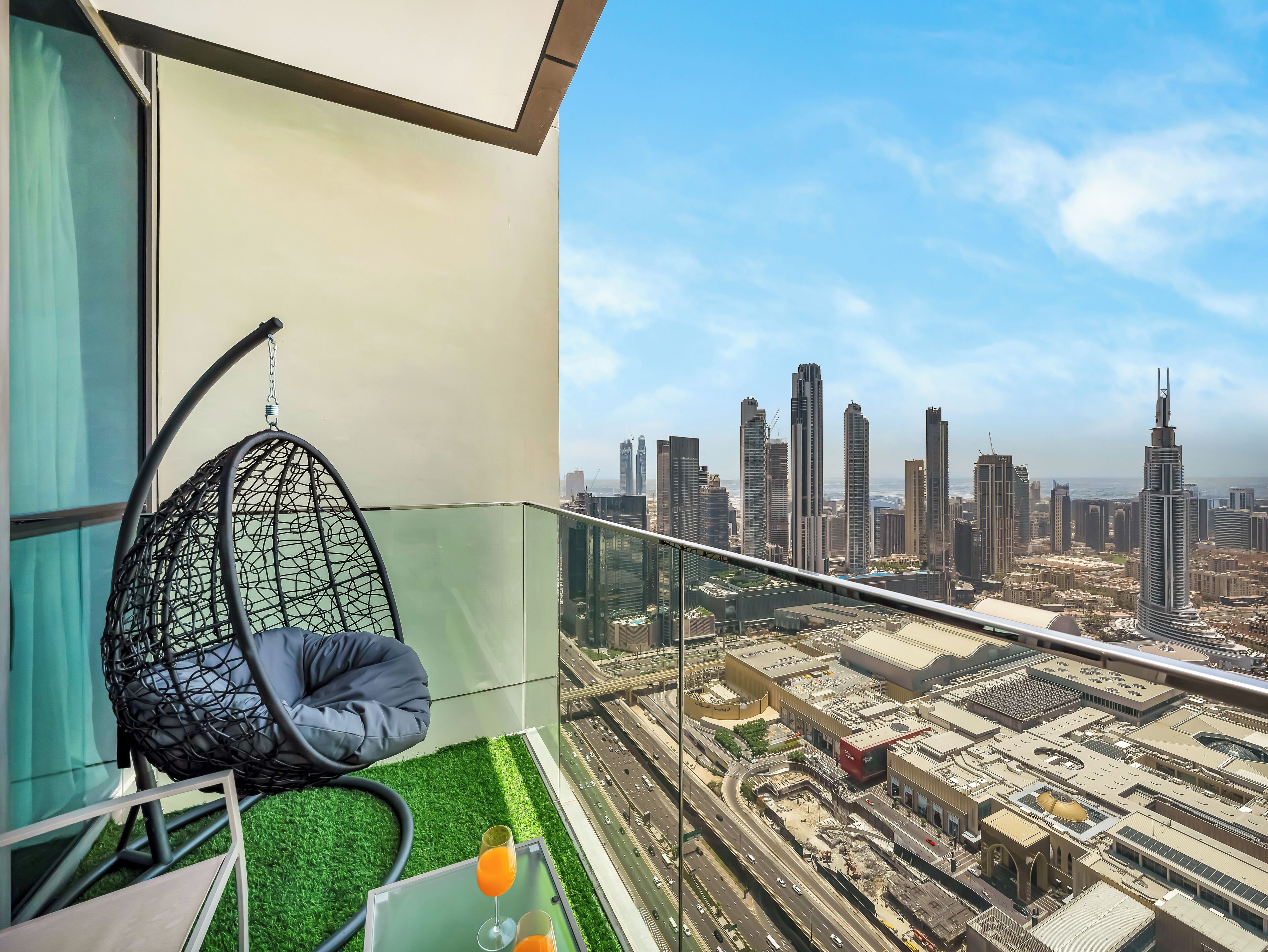 A balcony with a hanging chair, a glass table with drinks, and a cityscape view.