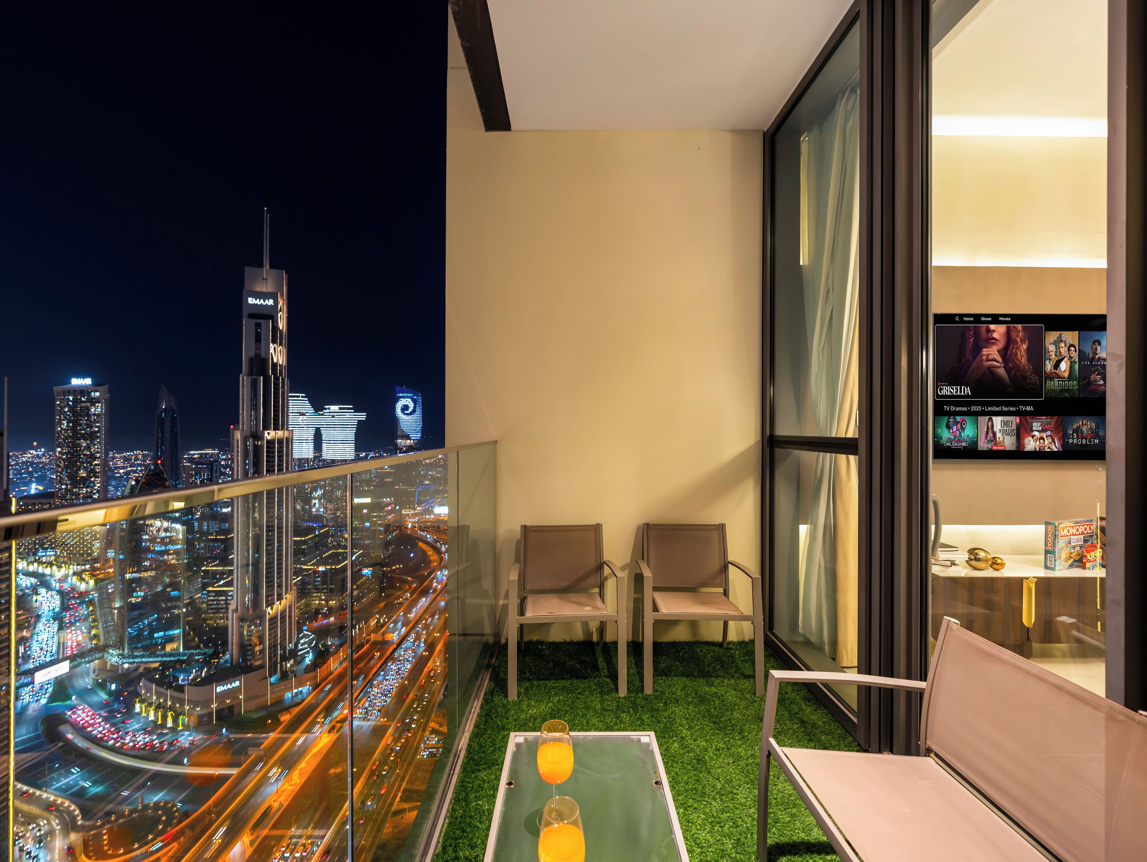 A balcony with a city view, two chairs, a small table with drinks, and a TV displaying a movie.