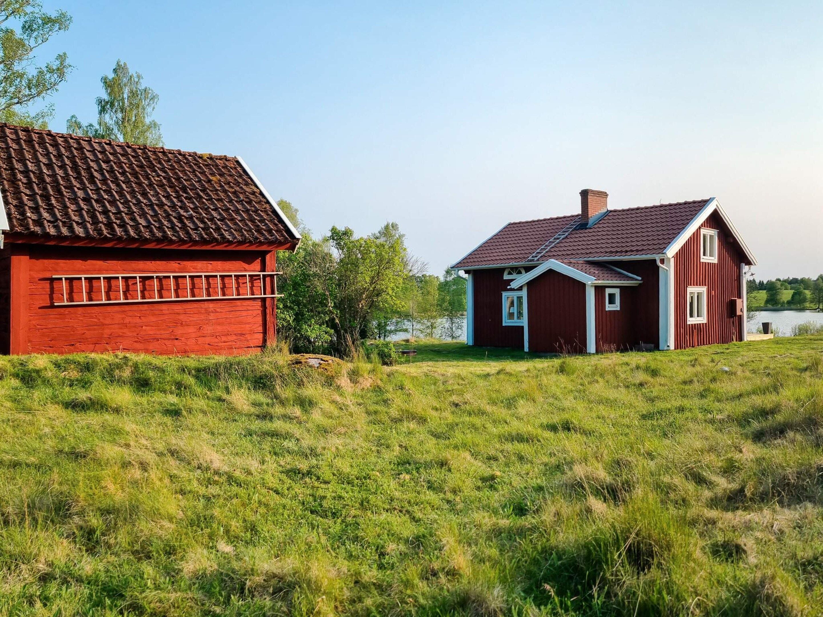 A red wooden house with a tiled roof, situated on a grassy field with a body of water in the background.