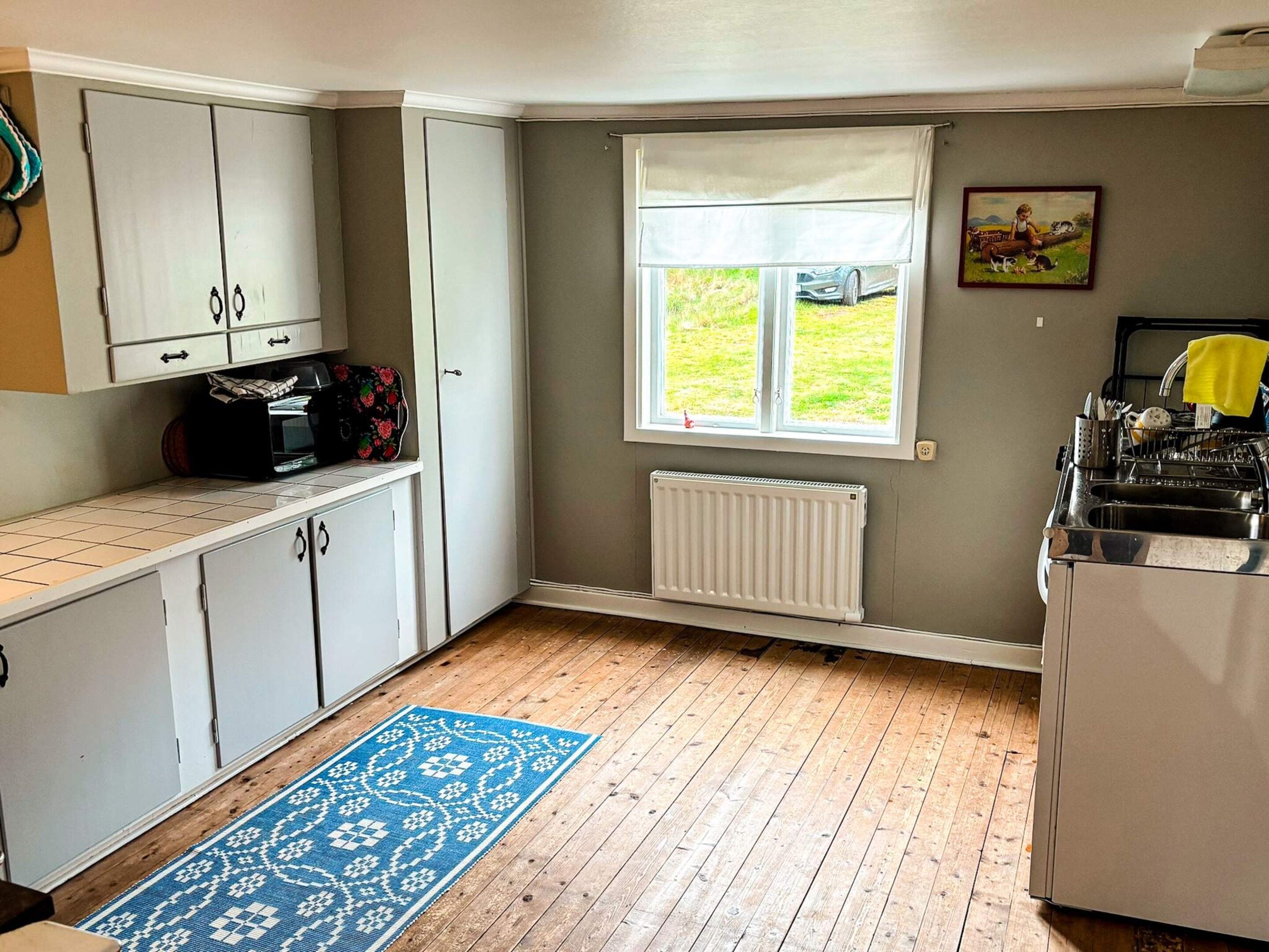 A kitchen with white cabinets, a window with a view, a radiator, and a blue patterned rug.