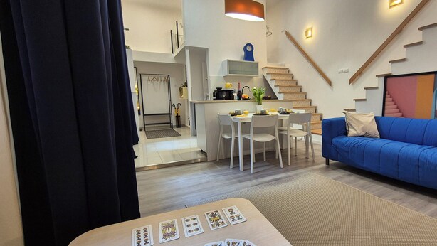 A modern living room with a blue sofa, a dining area with a table and chairs, a kitchen with a stove and sink, and a staircase with a colorful mural.