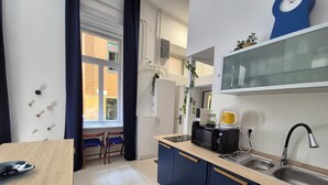 A modern kitchen with a microwave, kettle, and sink. There is a window with curtains, a plant, and a clock on the wall.