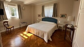 A bedroom with a wooden floor, a bed with a blue headboard, two bedside tables with lamps, a chair, and a window with curtains. - Historic house with garden across from NYU Florence (Unknown)