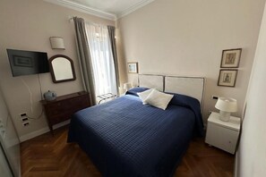 A bedroom with a bed, a dresser, a TV mounted on the wall, a nightstand with a lamp, and a window with curtains.