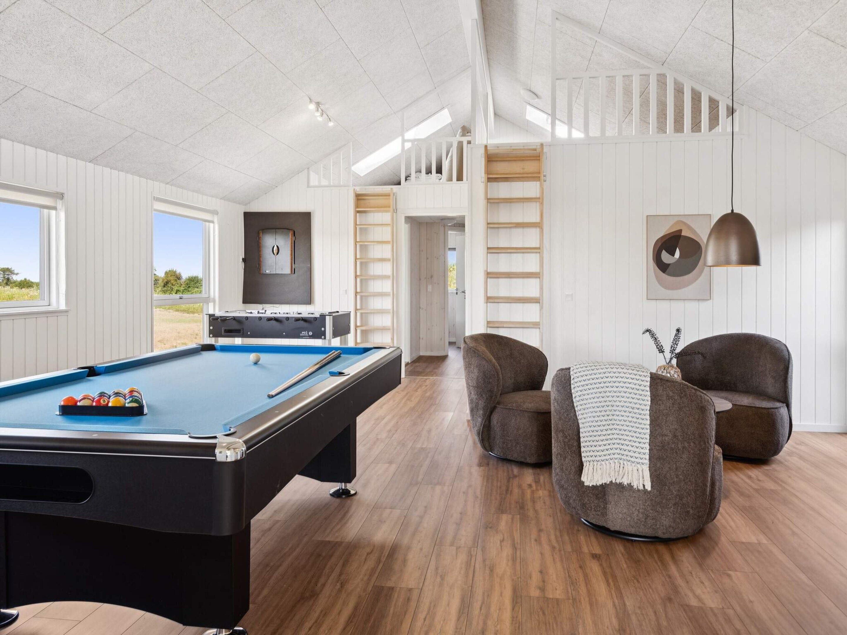 Une table de billard avec des billes, une queue de billard et une table de baby-foot dans une pièce au plancher en bois et aux murs blancs.