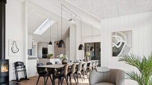 A modern living room with a fireplace, a dining area with a long table and chairs, and a kitchen in the background. - 5 Star Holiday Home in Bogense (Bogense)