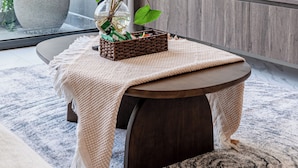 A modern living room with a round wooden table, a potted plant, a framed picture, and a large window with curtains.