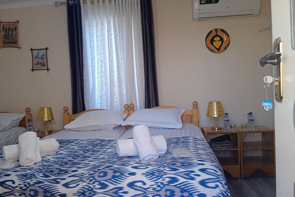 A hotel room with a bed, two bedside tables, a water dispenser, and a wall-mounted air conditioner.