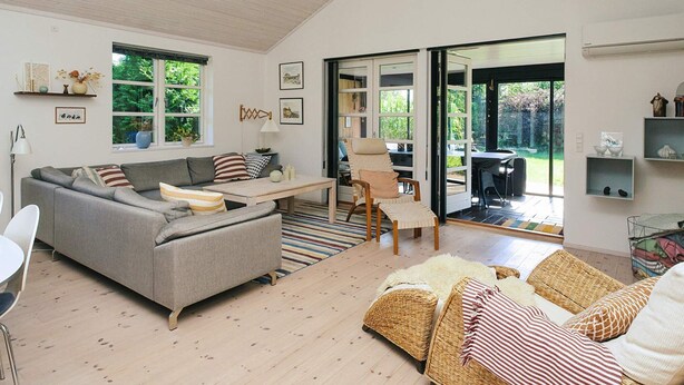 Ein geräumiges Wohnzimmer mit grauem Sofa, einem Holztisch, Korbmöbeln und Blick durch eine Glastür auf einen Außenbereich.