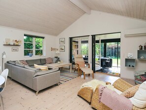 A spacious living room with a grey sofa, a wooden coffee table, wicker furniture, and a view through a glass door to an outdoor area.