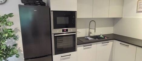 Una cocina moderna con horno empotrado, microondas y refrigerador. Hay un lavamanos con grifo y armarios con pomos. Un reloj se ve en la pared.