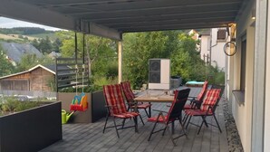 A covered patio with a table and chairs, a view of the surrounding landscape, and a modern building in the background.