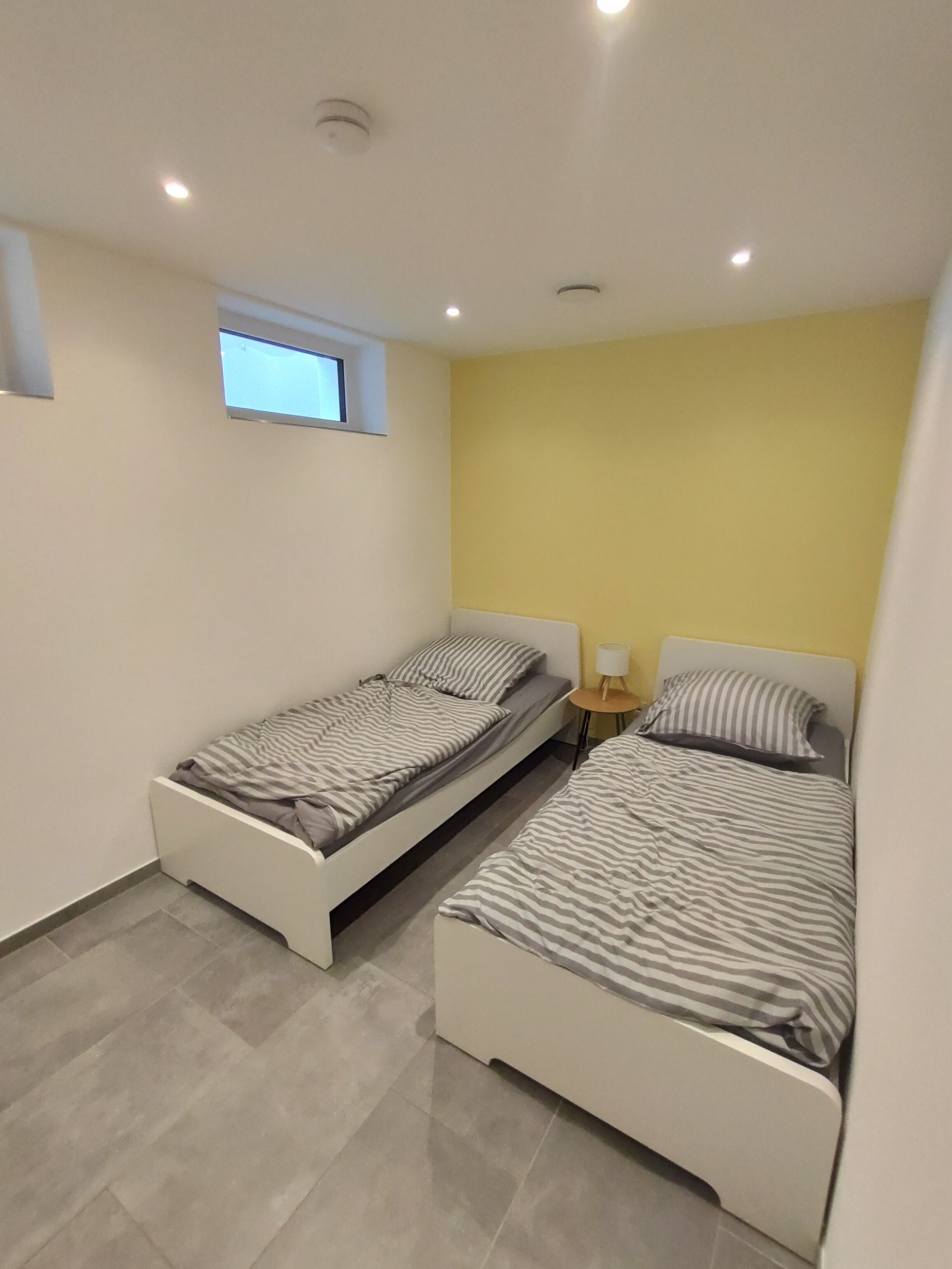 A small, modern bedroom with two single beds, a small window, and a yellow accent wall.