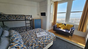Ein Etagenbett mit gemusterten Bettbezügen, ein Sofa mit gelben Kissen, ein Fernseher an der Wand und ein Fenster mit Aussicht.