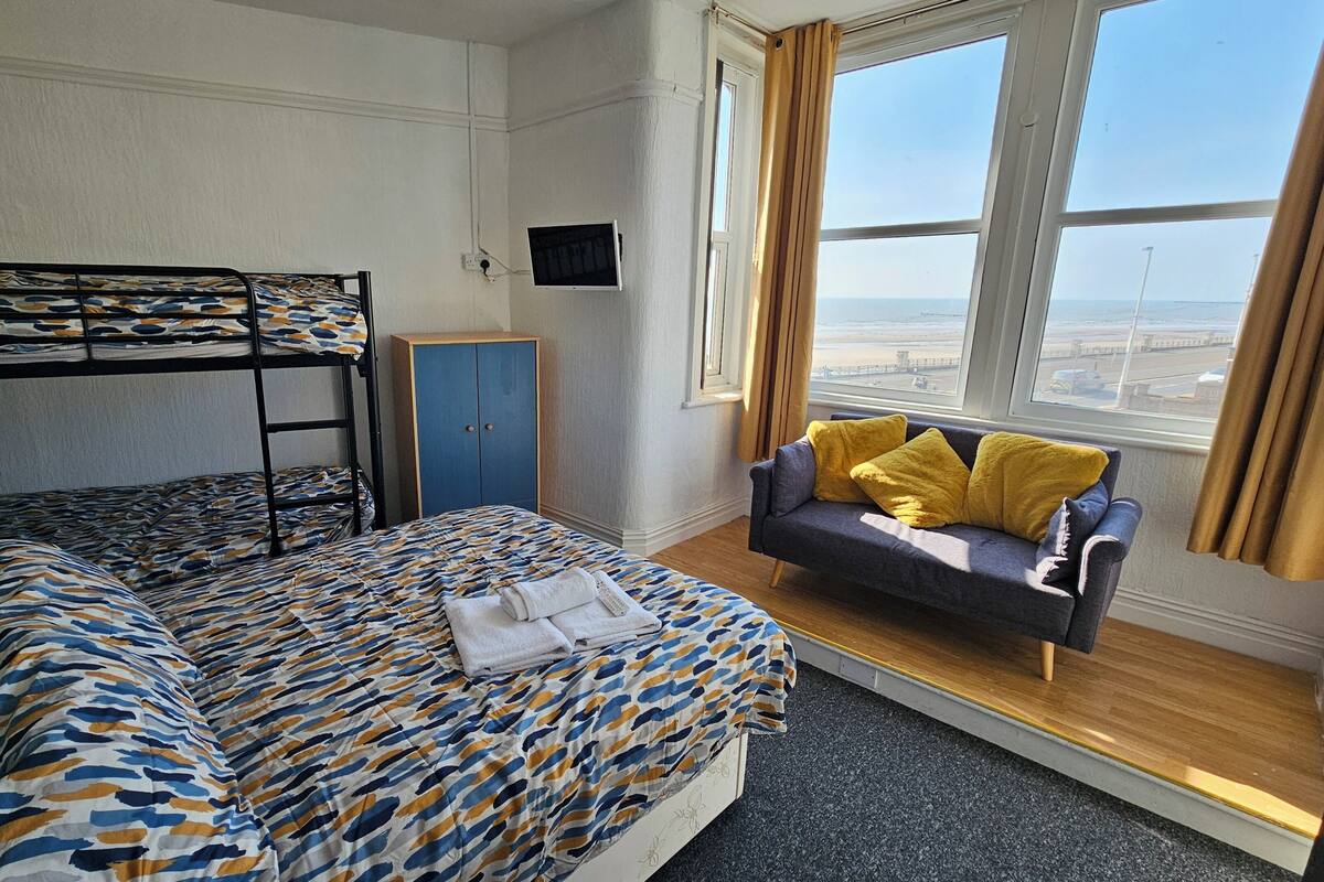 Una cama litera con ropa de cama estampada, un sofá con cojines amarillos, un televisor en la pared y una ventana con vistas.