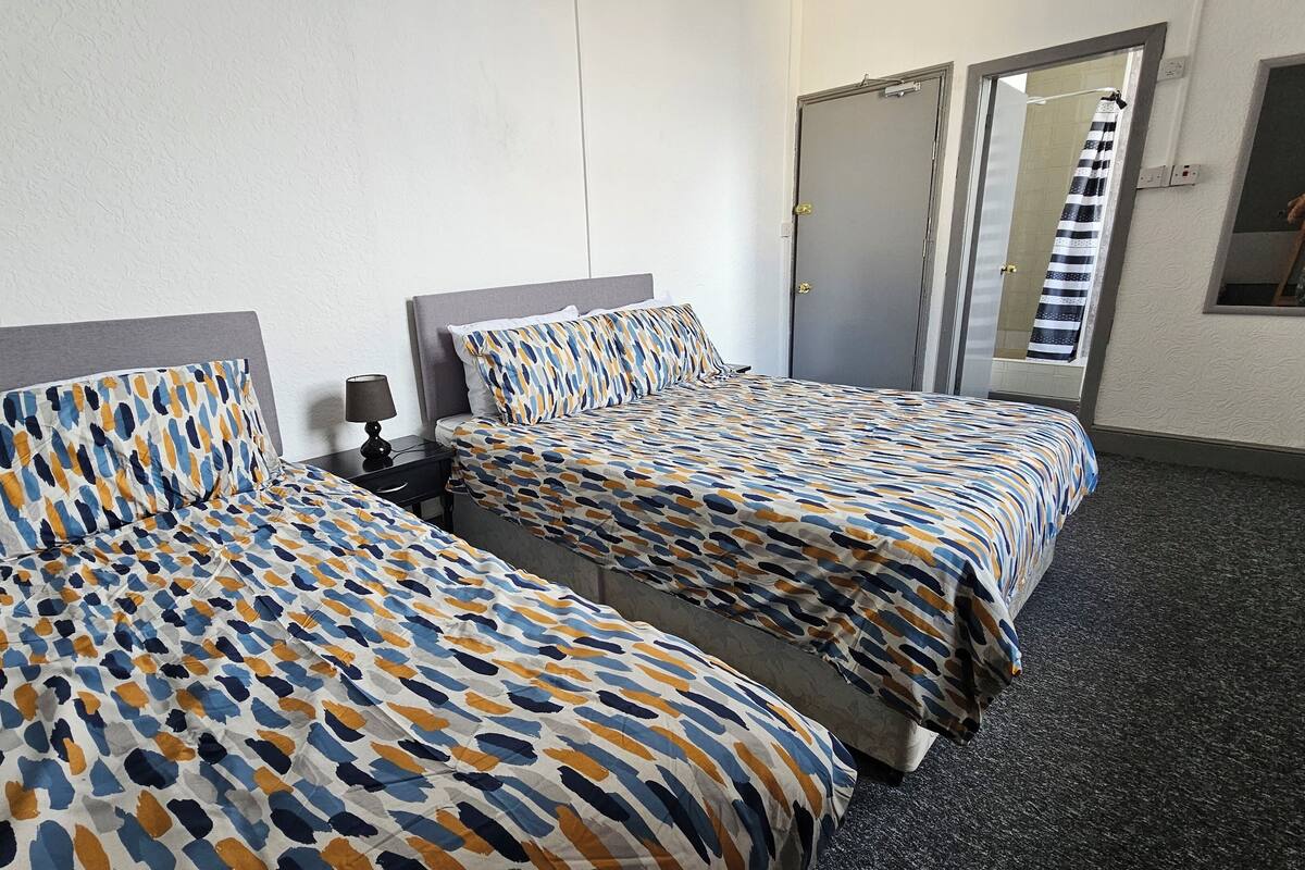 Habitación de hotel con dos camas, cada una con ropa de cama estampada, una mesita de noche con lámpara y una puerta con una toalla colgada.